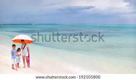Family walking with colorful orange umbrella on a white sand tropical beach at rainy day
