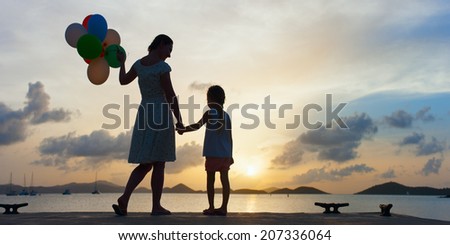 Silhouette of happy mother and her little daughter with bunch of balloons on sea coast at sunset