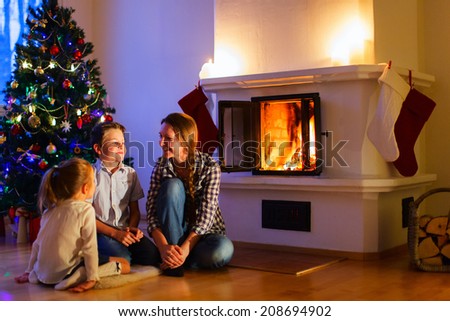 Mother and her two little kids sitting by a fireplace in their cozy family home enjoying winter evening