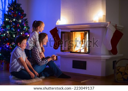 Mother and her two little kids sitting by a fireplace in their family home on Christmas eve