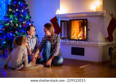 Family of mother and her two little kids sitting by a fireplace in their home on Christmas eve