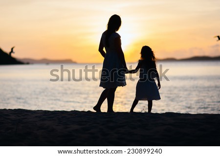 Silhouettes of mother and daughter walking along tropical beach during sunset