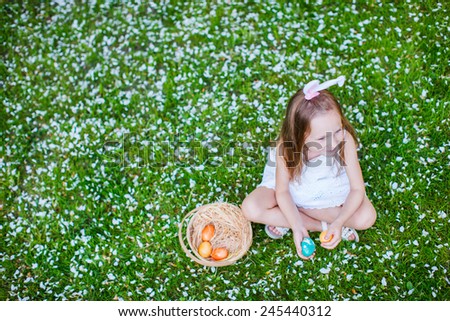 Top view of adorable little girl wearing bunny ears playing with Easter eggs on a grass covered with white flower petals on spring day