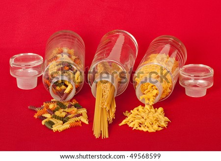 Pasta jars with macaroni, spaghetti, and egg noodles spilled onto a red table cloth