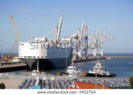 A large car carrier ship moored in the harbor with cars in the foreground waiting to be loaded onboard