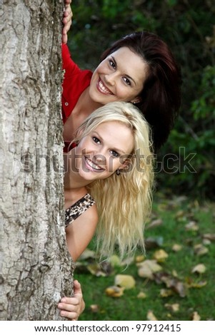 Two lovely young smiling women peeping out from behind a tree trunk