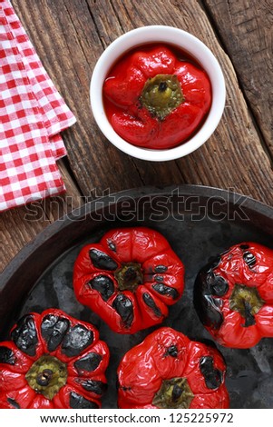 Pickled red pepper: roasted and skinned red pepper marinated in a sauce of vinegar and olive oil, next to a pan with freshly roasted peppers. Rustic take on an old wooden table.