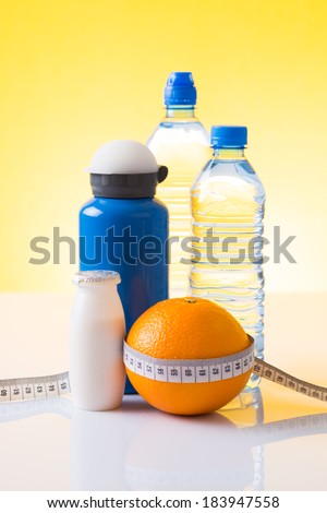 Measuring tape wrapped around an orange, bottled water and blue sport bottle as a symbol of diet on yellow background.