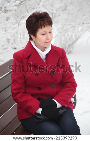 Attractive elderly woman in red coat sitting and resting on bench in winter snowy street