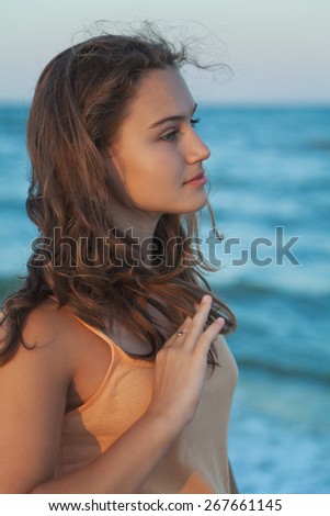 Young long-haired teen girl with brown hair standing in profile on the beach and watching to sea on a summer evening