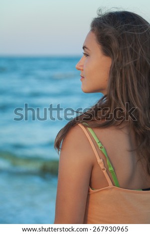 Young long-haired teen girl with brown hair standing in profile on the beach and watching to sea on a summer evening