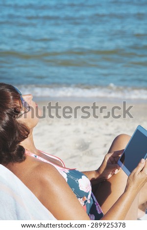 Woman with tablet pc at the beach. Elderly woman in a swimsuit sunbathing and holding a tablet on summer sea beach. Woman looks up and enjoy vacation at sea.