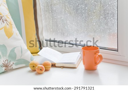 Three apricots on windowsill at the window with raindrops next to the book, pillow and cup on a rainy day. Set for a cozy summer reading.