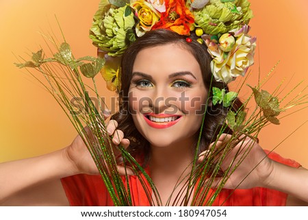 Beautiful smiling girl wearing flower headband looking through grass with green butterflies over orange background