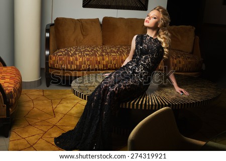 Beautiful young woman with blonde hair dressed in long lace dress sitting on round table.