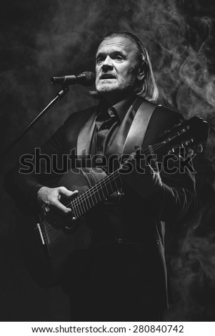 Old man with grey hair playing guitar and singing over smoky black background