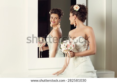 Beautiful young bride in stylish wedding gown standing near mirror