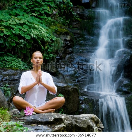 Beautiful young woman meditating in lotus position while doing yoga in a wonderful forest near waterfall