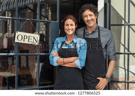 Photo of Two cheerful small business owners smiling and looking at camera while standing at entrance door. Happy mature man and mid woman at entrance of newly opened restaurant with open sign board.