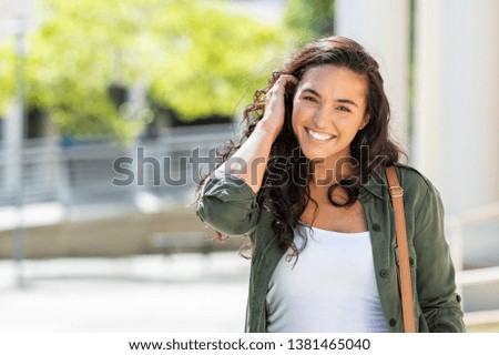 Happy young beautiful woman walking on the street. Portrait of cheerful university student looking at camera while adjusting curly hair with copy space. Latin stylish girl smiling while standing. 商業照片 © 