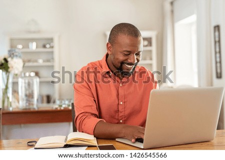 Photo of Smiling black man using laptop at home in living room. Happy mature businessman send email and working at home. African american freelancer typing on computer with paperworks and documents on table.