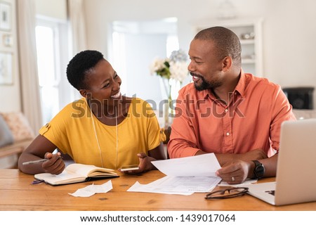 Mature couple sitting and managing expenses at home. Happy african man and woman paying bills and managing budget. Black smiling couple checking accountancy and bills while looking at each other.