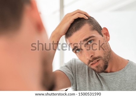 Young unshaven man looking at mirror in bathroom at home. Handsome guy looking at his face in mirror, checking hair and hairline. Man in pijamas concerned with hair loss.
