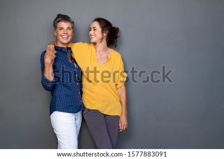 Mature happy women embracing each other against grey wall with copy space. Happy laughing ladies standing on gray background. Cheerful middle aged woman with hand on shoulder of her stylish friend.