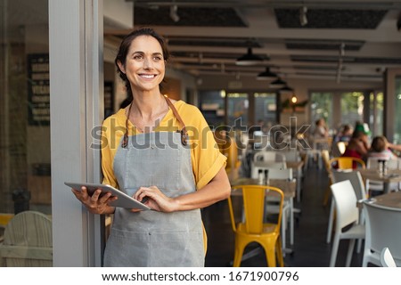 Portrait of a happy waitress standing at restaurant entrance holding digital tablet. Happy mature woman owner in grey apron standing at coffee shop entrance leaning while looking away with copy space.