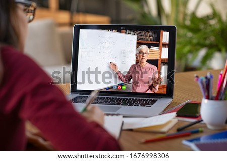 Young student watching lesson online and studying from home. Young woman taking notes while looking at computer screen following professor doing math on video call. Girl studying from home on pc.