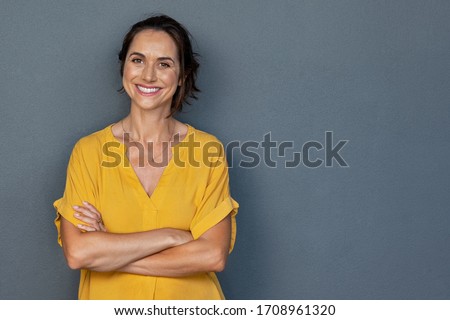 Confident mature woman with crossed arms in casual clothing with copy space. Successful smiling woman with big grin looking at camera. Beautiful positive businesswoman standing against grey background