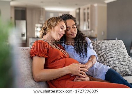 Multiethnic lesbian couple with hands on baby bump of pregnant woman. Expecting mother feeling baby kick with her best friend sitting on couch. Young mixed race woman touching belly of her girlfriend.