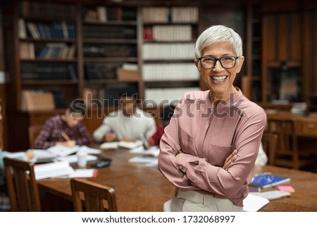 Portrait of mature professor standing in university library and looking at camera with copy space. Happy senior woman at the library working as a librarian. Satisfied college teacher smiling.