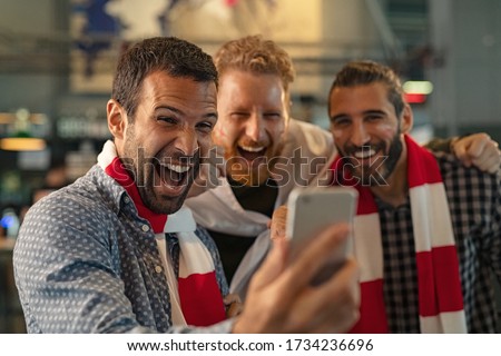 Excited men watching football in streaming on smartphone. Football fans watching game on phone and celebrating victory score at pub. Happy supporters cheering and exulting after winning an online bet.