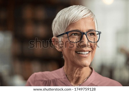Portrait of smiling mature woman with spectacles in library looking away. Senior librarian standing in reading hall and thinking. Old beautiful lecturer contemplating. Future and vision concept.