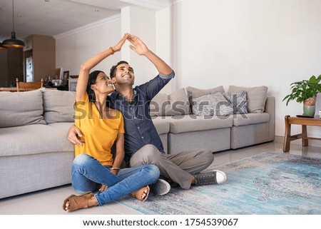 Handsome young man with beautiful indian woman dreaming a new home. Smiling married couple moves to new apartment with copy space. Happy middle eastern couple making roof with hands symbol of new home