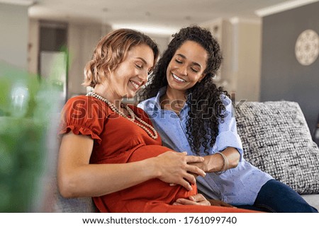 Lovely young lesbian couple planning the future of their baby. Happy mixed race woman smiling with hands on expecting mother's baby bump while feeling baby movement; assisted fertilization and ivf.