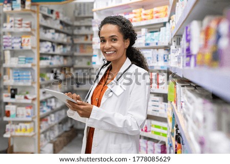 Happy friendly multiethnic pharmacist doing inventory in a provided and modern pharmacy while looking at camera. Portrait of smiling african doctor woman working in drugstore with digital tablet.