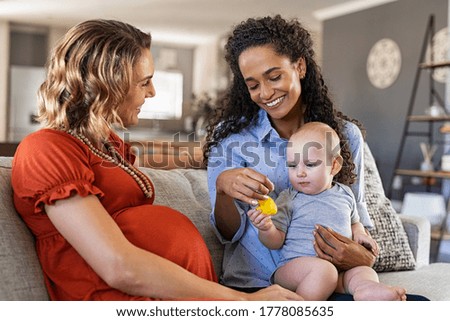 Happy multiethnic friends with child sitting on couch. Pregnant mother relaxing with her girlfriend. Pregnant lesbian gay couple with one toddler son, assisted fertilization and adoption concept. 