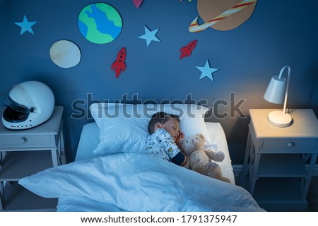 High angle view of little boy dreaming of becoming an astronaut while sleeping with teddy bear in space decorated room. Top view of dreamer child sleeping on bed during the night and the light on.