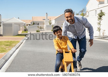 Happy latin father helping smiling boy to ride wooden balance cycle on street. Happy middle eastern child and young dad riding bike. Smiling daddy teaching son to ride a balance bicycle, copy space.