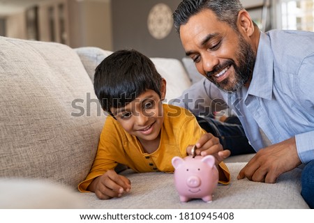 Happy indian son saving money in piggy bank with father. Lovely ethnic father teaching to little boy importance of saving money for future. Smiling middle eastern kid adding coin in piggybank with dad