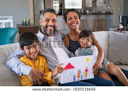 Proud parents showing family painting of son and daughter sitting on sofa at home. Smiling african mother and middle eastern father with two children looking at camera. Brothers showing painting.