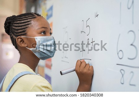 Portrait of african girl wearing face mask and writing solution of sums on white board at school. Black schoolgirl solving addition sum on white board during Covid-19 pandemic. School child thinking. 商業照片 © 