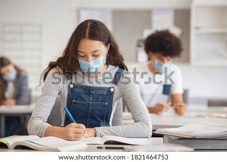 High school student taking notes while wearing face mask due to coronavirus emergency. Young woman sitting in class with their classmates and wearing surgical mask due to Covid-19 pandemic.