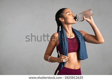 Fitness woman drinking water standing on gray background with copy space. Portrait of sweaty latin woman take a break after intense workout. Mid adult lady drink from water bottle after gym workout. 