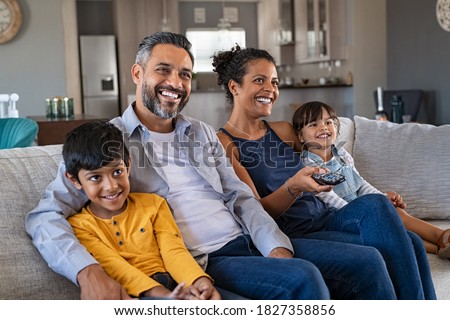 Happy indian man and african woman relaxing with daughter and son watching television at home together. Cheerful ethnic family relaxing on sofa at home watching movie with children. 