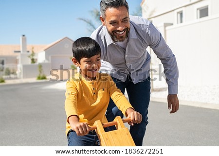 Cheerful middle eastern father helping excited son to ride wooden balance cycle on street. Happy boy enjoying riding bycicle with his dad. Dad teaching his indian son to ride bicycle outdoor.