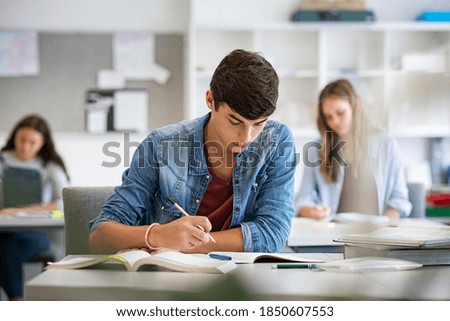 Focused young man taking notes from books for his study. College student sitting at desk and studying in high school library. Guy studying in classroom and completing project.