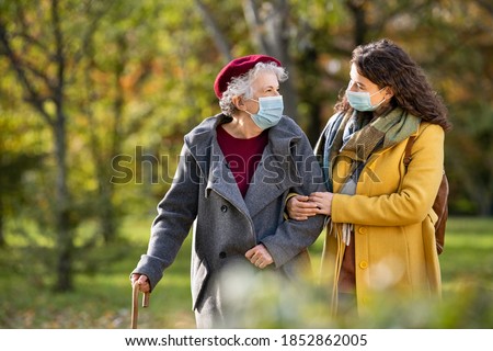 Lovely granddaughter walking with senior woman in park and wearing face mas against covid-19. Smiling old grandmother with happy caregiver in park relaxing after quarantine due to coronavirus.